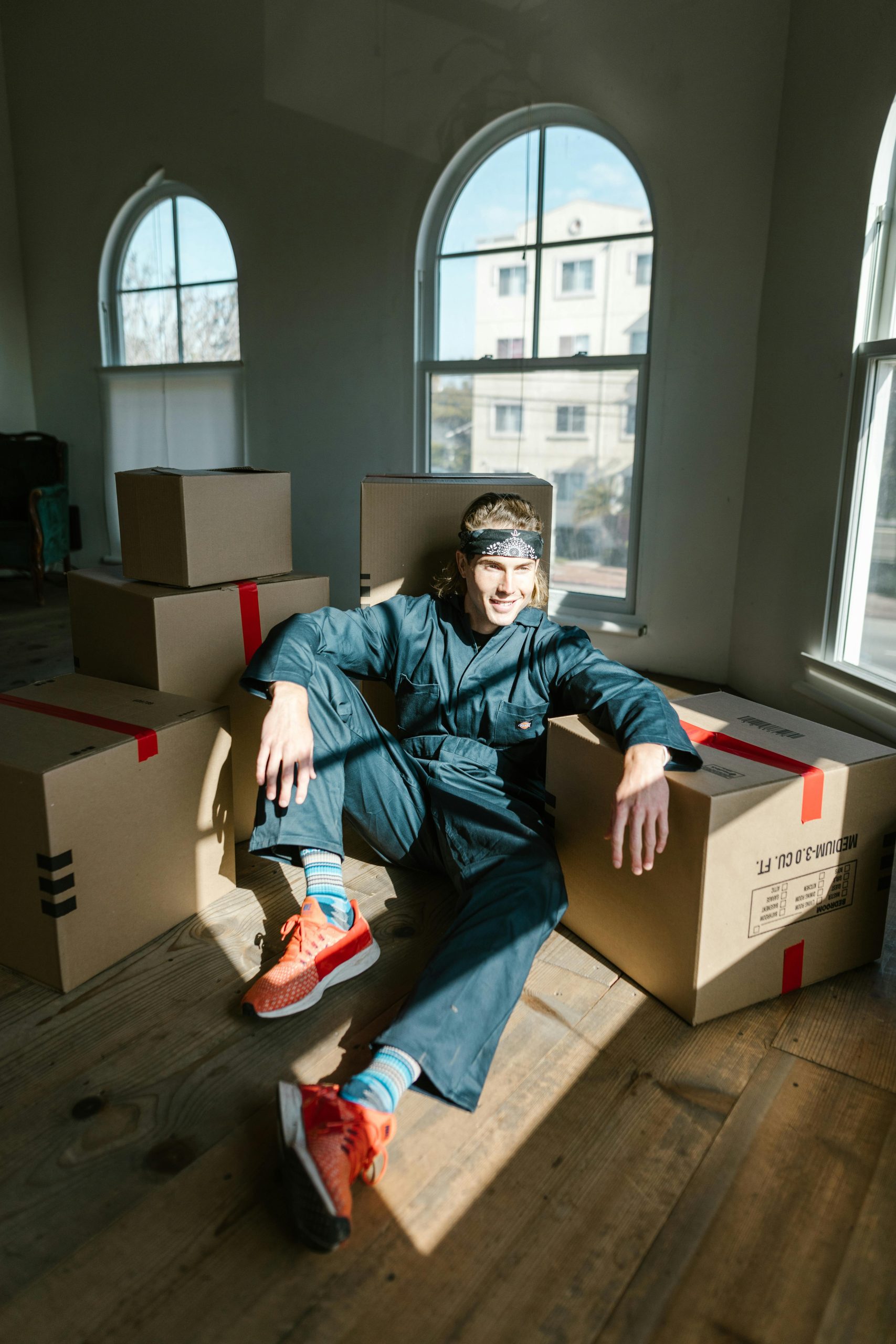 Man sitting among moving boxes in a sunlit room, wearing coveralls and looking relaxed.